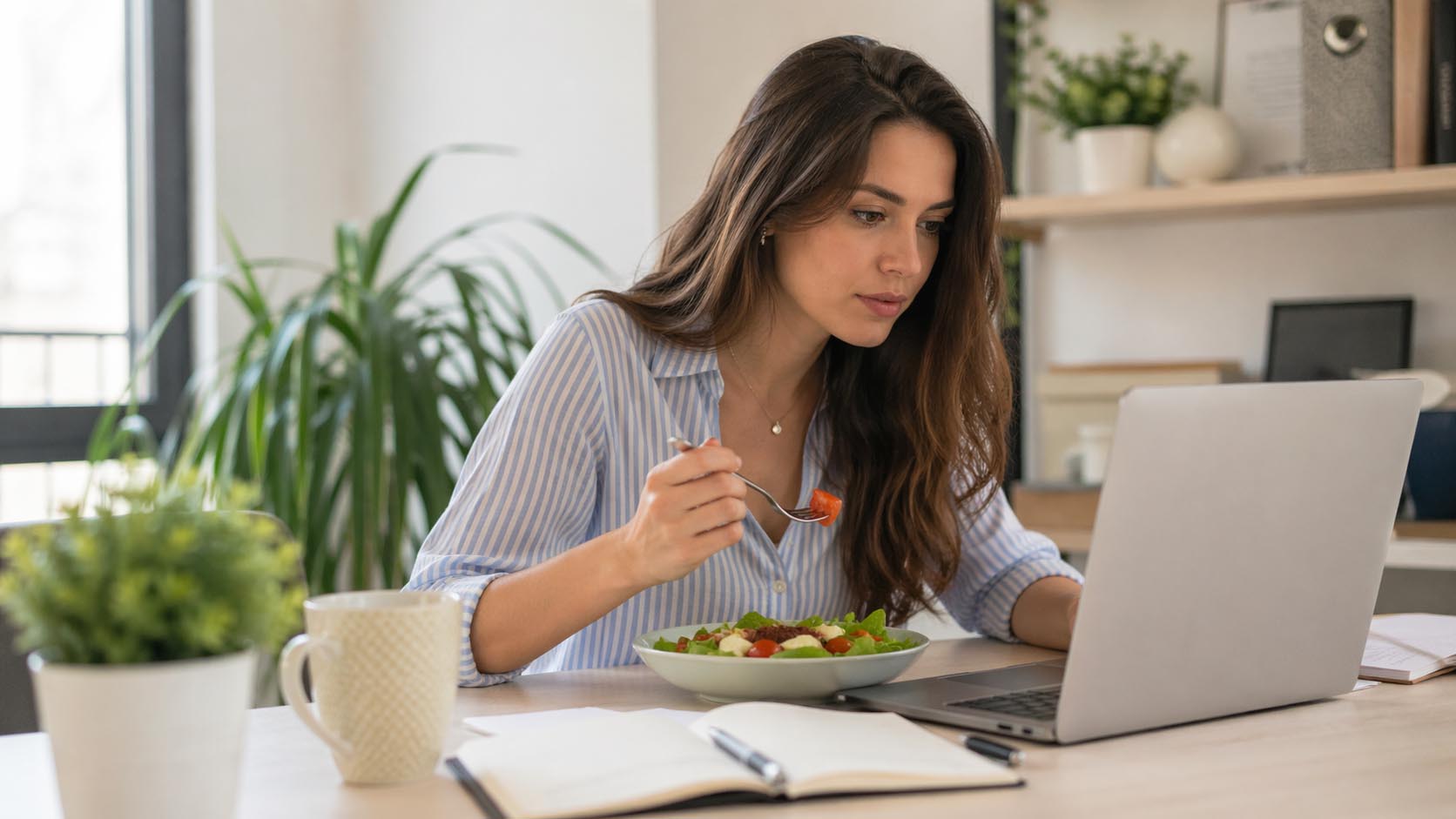 Mlada osoba jede lagan obrok i radi fokusirano za laptopom.